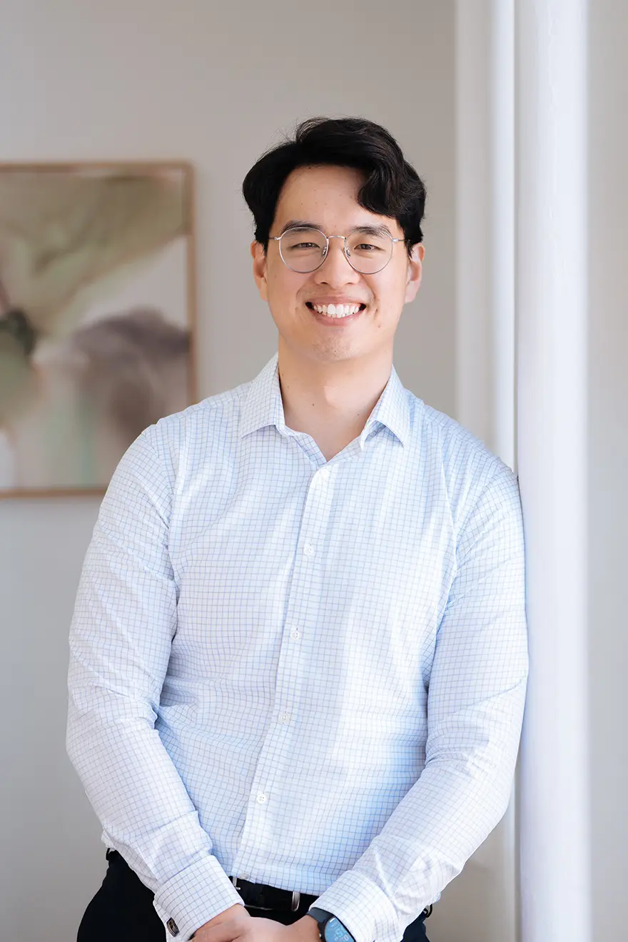 Kenny_Chong Kenny Chong, Senior Accountant, wearing a white collared shirt, smiling while leaning against a wall of a light-filled, elegant room