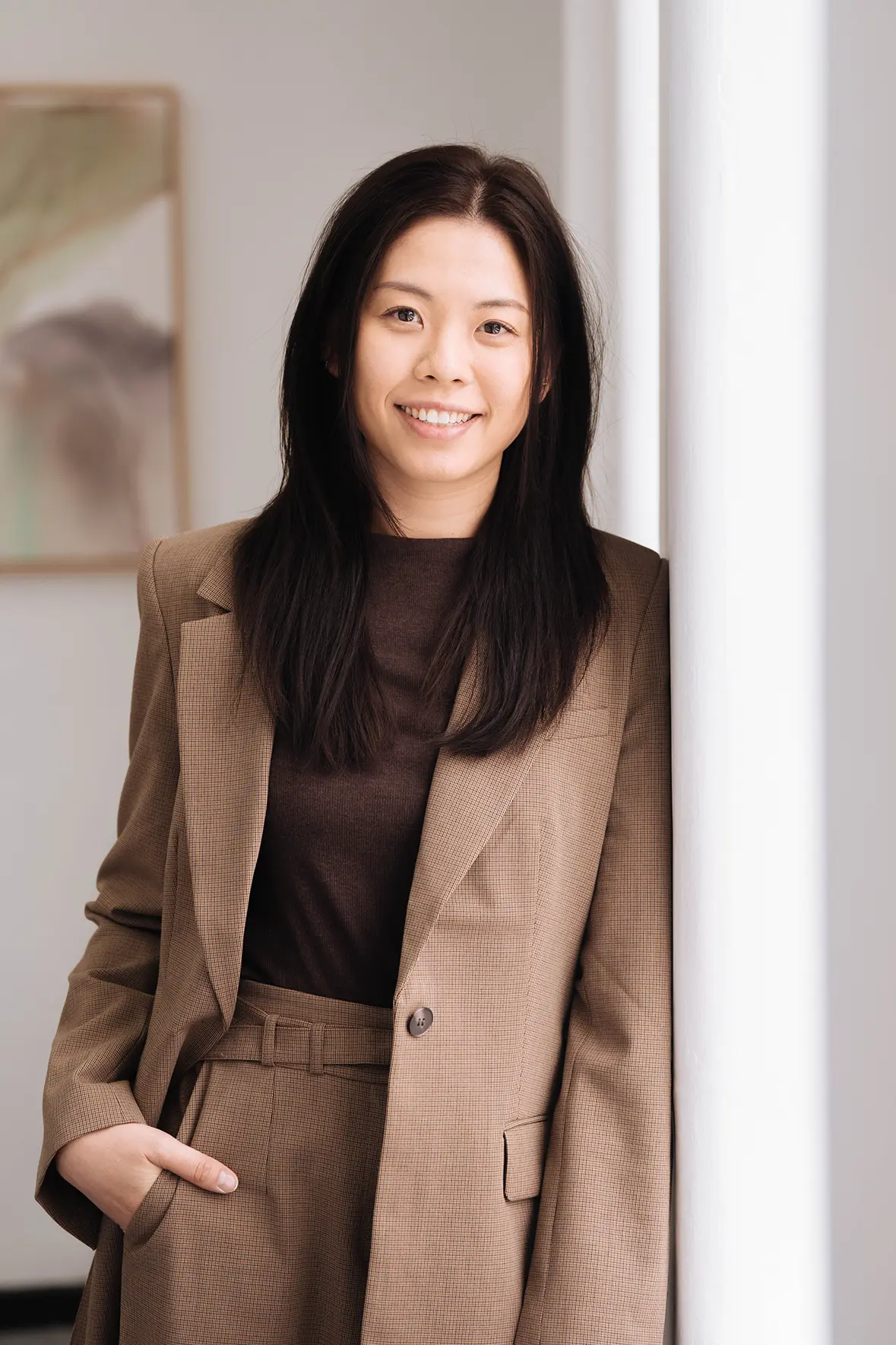 Rinni_Au Rinni Au, Senior Accountant, wearing dark brown business attire, smiling while leaning against a wall of a light-filled, elegant room